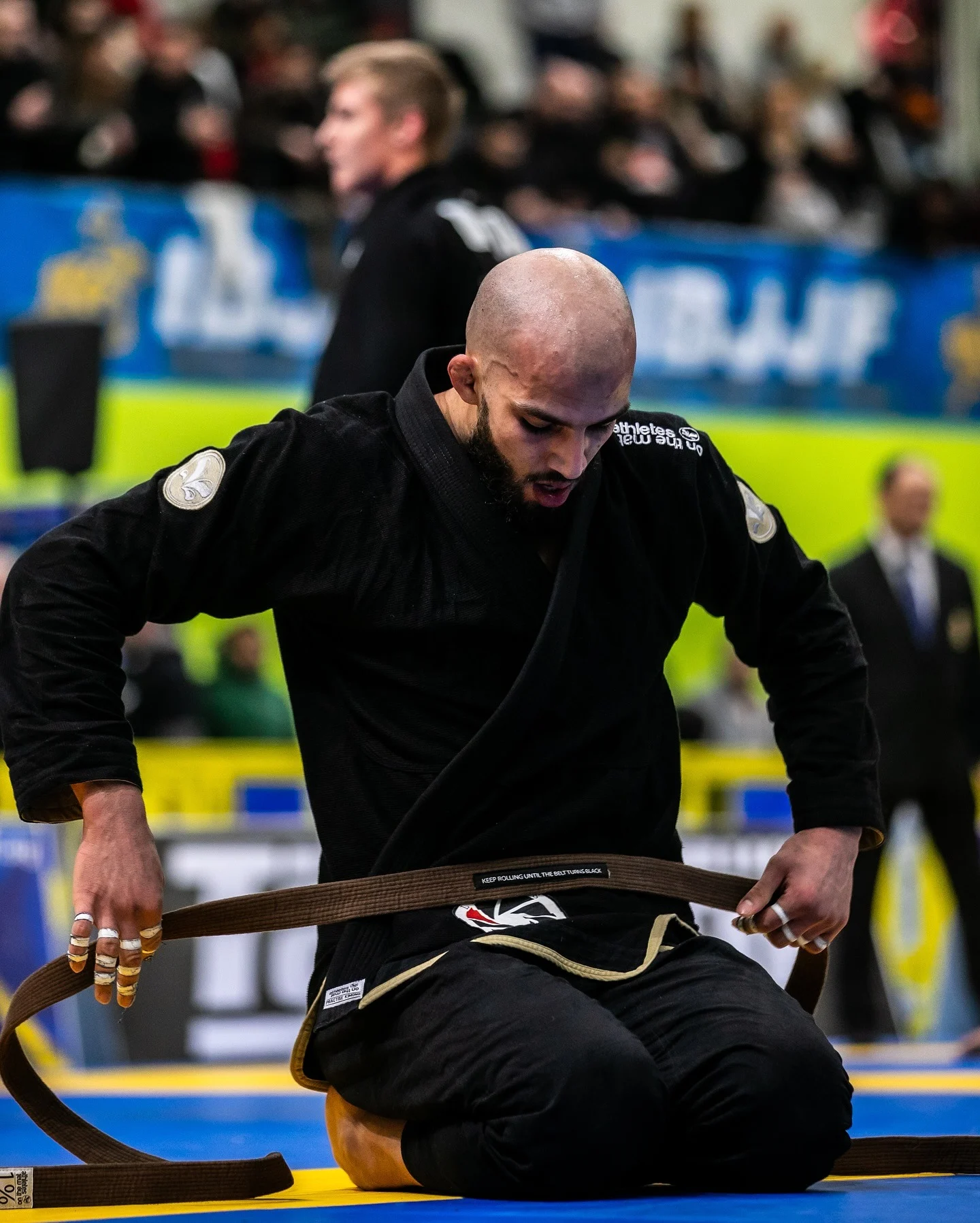 Reda Hamzaoui à genoux sur le tapis d'un championnat IBJJF, il noue sa ceinture marron