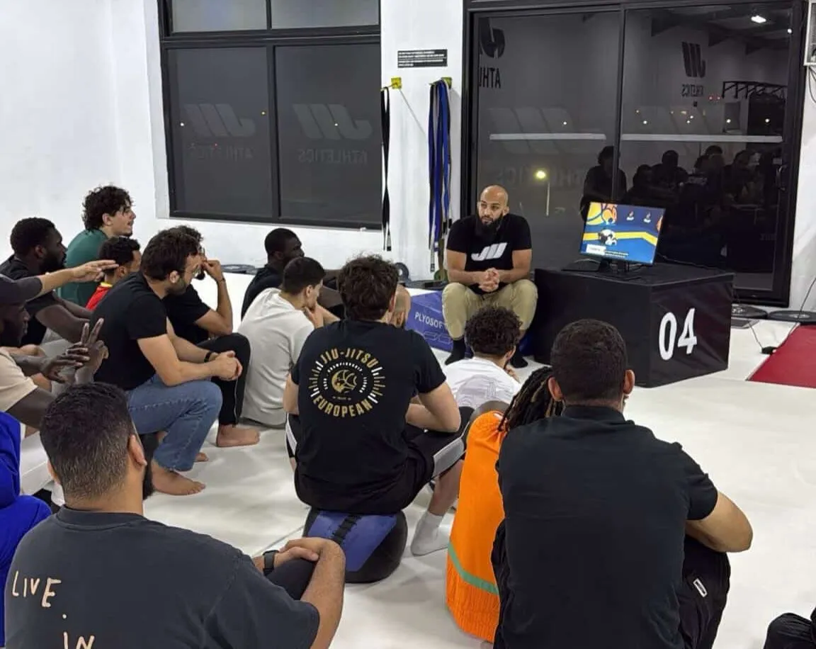 Reda commente un combat sur écran devant des athlètes assis sur le tapis